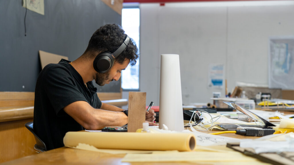 A designer wearing headphones works at a desk with various design tools and materials