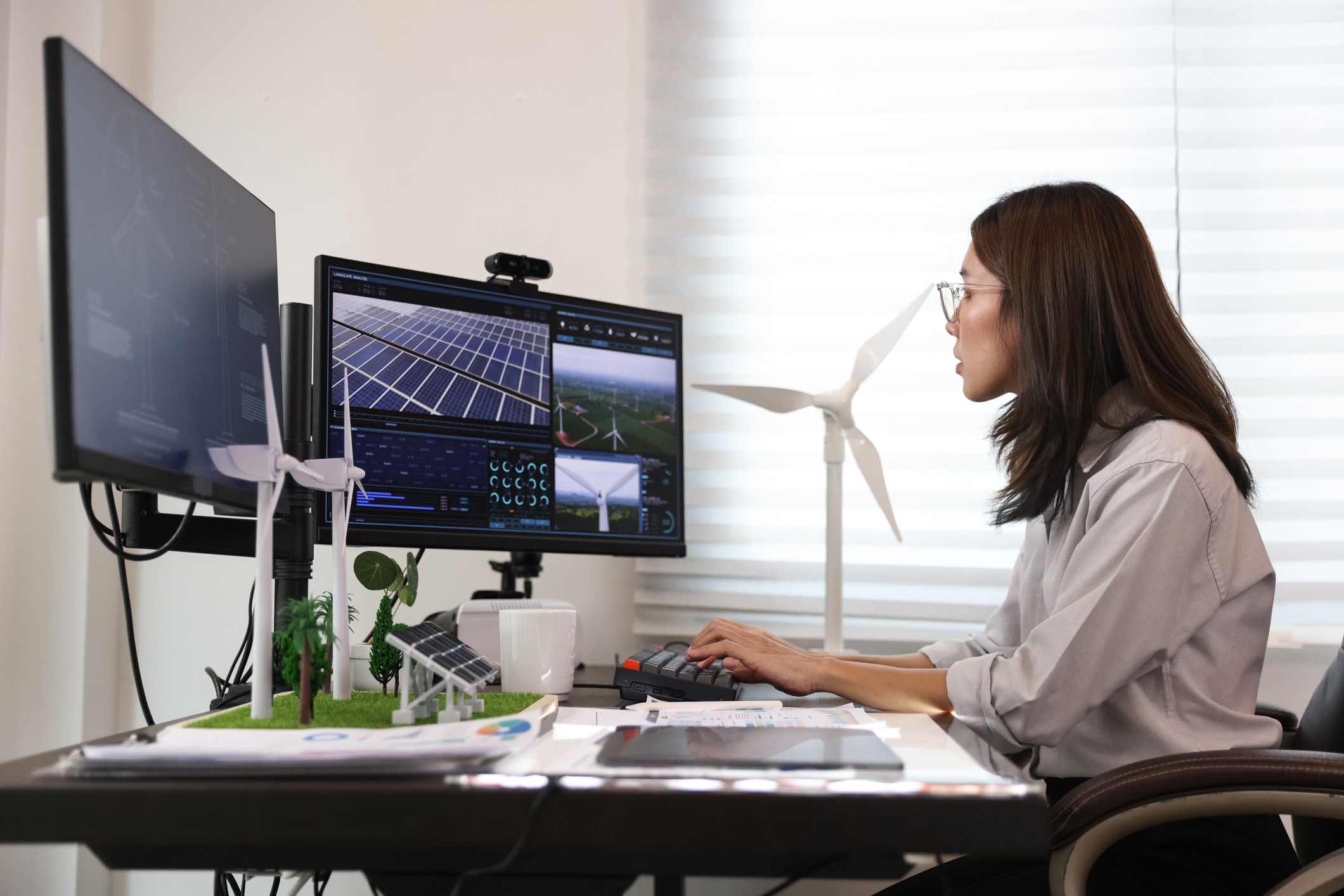 A woman works on a computer at a desk with model of renewable energy equipment